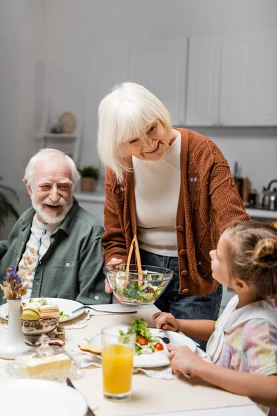 Усміхнена старша жінка тримає овочевий салат біля онуки під час великодньої вечері — стокове фото