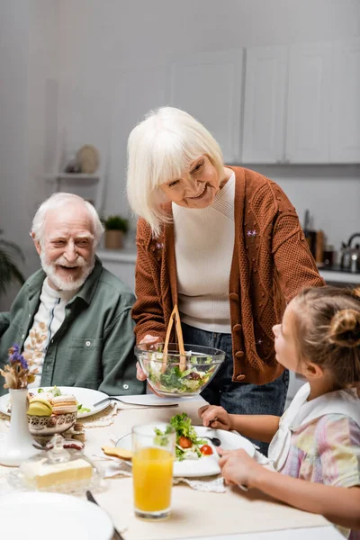 Mulher sênior sorrindo segurando salada de legumes perto da neta durante o jantar de Páscoa — Fotografia de Stock