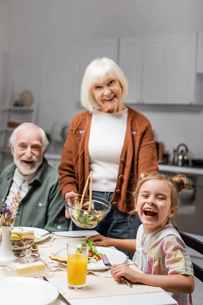 Femme âgée tenant salade de légumes près mari gai et petite-fille riante — Photo de stock