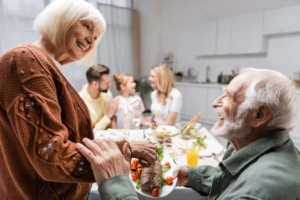 Lächelnde Frau mit gebratenem Fleisch neben älterem Mann beim Familienessen — Stockfoto