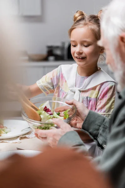 Chica sonriente sosteniendo ensalada de verduras cerca del abuelo sobre fondo borroso - foto de stock