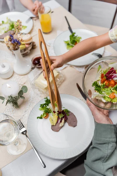 Vista recortada de chica con pinzas de servir poniendo ensalada de verduras en el plato durante la cena de Pascua — Stock Photo