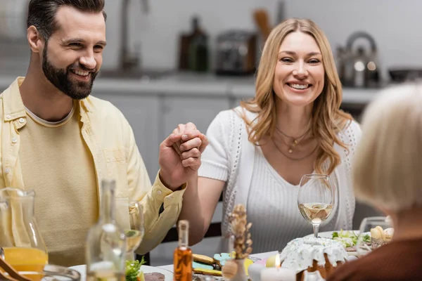 Allegra coppia che si tiene per mano vicino al tavolo servito con cena pasquale in primo piano sfocato — Foto stock