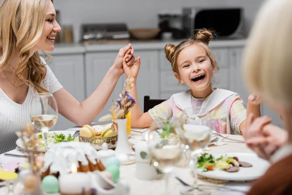 Chica sorprendida de la mano con la madre y la abuela cerca de la cena festiva en la mesa de la cocina - foto de stock