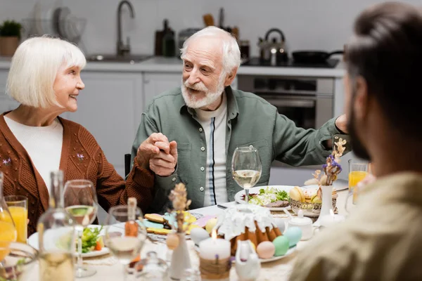 Glückliches Senioren-Paar Händchen haltend beim Osteressen mit verschwommenem Sohn — Stockfoto