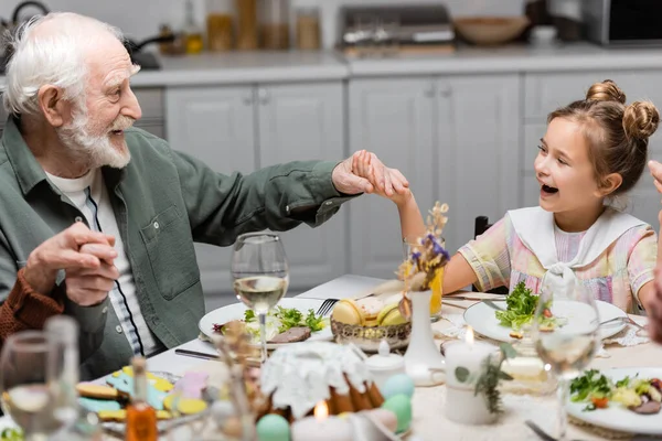 Excitada chica cogida de la mano con el abuelo feliz durante la cena de Pascua en casa - foto de stock