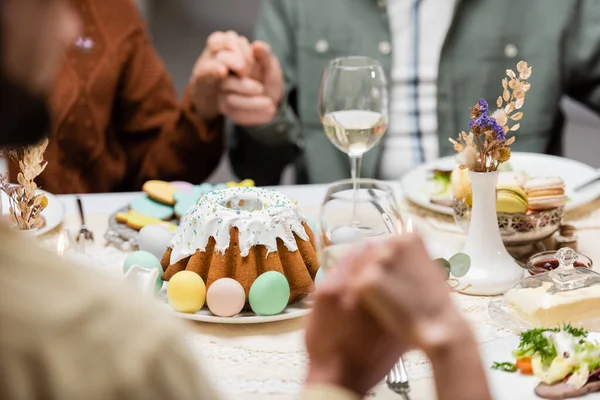 Vista recortada de la familia cogida de la mano cerca de pastel de Pascua y huevos pintados en la mesa - foto de stock