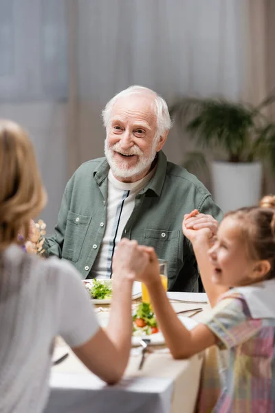 Старший мужчина улыбается рядом с женщиной с дочерью, держась за руки во время пасхального ужина — стоковое фото