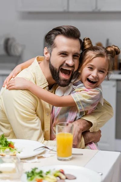 Aufgeregter Mann und Tochter umarmen sich nahe Glas Orangensaft auf Tisch — Stockfoto