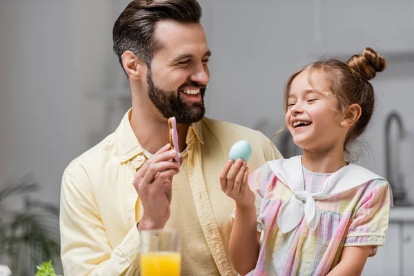 Alegre hombre e hija sosteniendo galleta y huevo pintado durante la celebración de Pascua - foto de stock