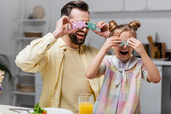 Веселий чоловік з дочкою прикриває очі великодніми яйцями та печивом, розважаючись вдома — стокове фото