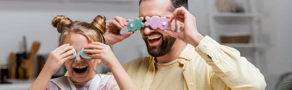 Lachende Vater und Tochter, die die Augen mit Osterkeksen und bemalten Eiern bedecken, während sie zu Hause Spaß haben, Banner — Stockfoto