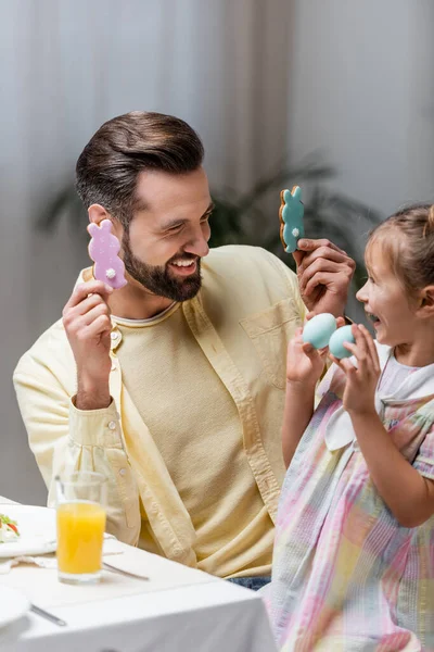 Excitada chica con papá divirtiéndose con huevos pintados y galletas tradicionales durante la cena de Pascua - foto de stock