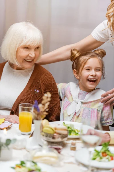 Fröhliches Mädchen lacht neben Oma und Mama am Tisch, serviert mit festlichem Abendessen — Stockfoto