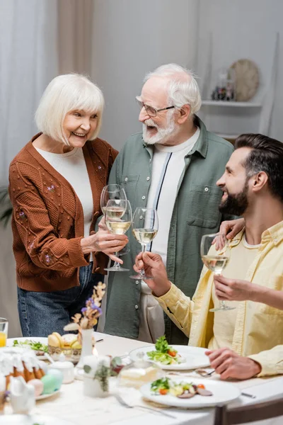 Glückliches Senior-Paar klimpert beim Osteressen mit erwachsenem Sohn Weingläser — Stockfoto