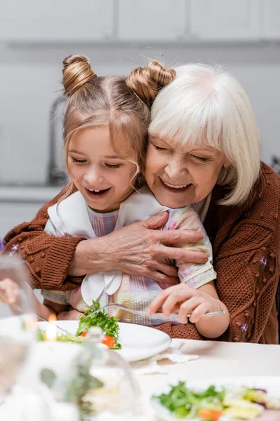 Glückliche Seniorin umarmt Enkelin, die beim Osteressen Gemüsesalat isst — Stockfoto