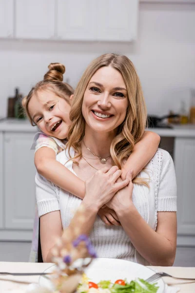 Cheerful girl embracing happy mother while looking at camera near served dinner — Stock Photo