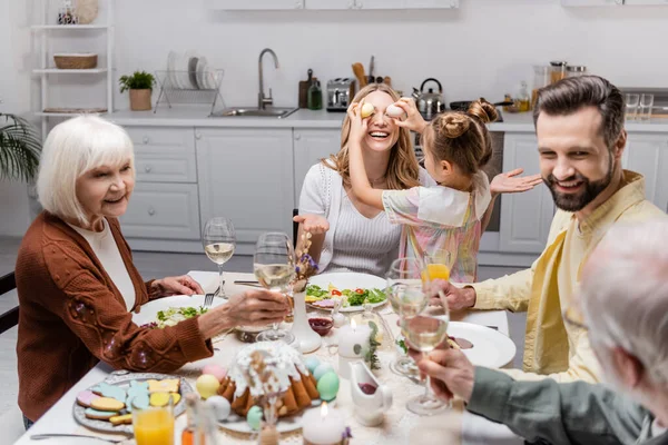 Ragazza che copre gli occhi della madre con uova di Pasqua mentre la famiglia brinda con bicchieri di vino — Foto stock