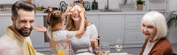 Chica cubriendo los ojos de feliz mamá mientras se divierten durante la cena de Pascua con la familia, pancarta - foto de stock