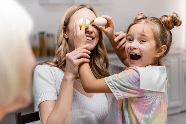 Excitada chica cubriendo los ojos de la madre con huevos de Pascua cerca borrosa abuelita - foto de stock