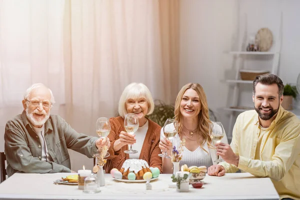 Familia complacida sosteniendo copas de vino y sonriendo a la cámara cerca de pastel de Pascua y huevos pintados - foto de stock
