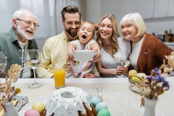 Chica emocionada con la boca abierta tomar selfie en el teléfono inteligente durante la cena de Pascua con la familia - foto de stock