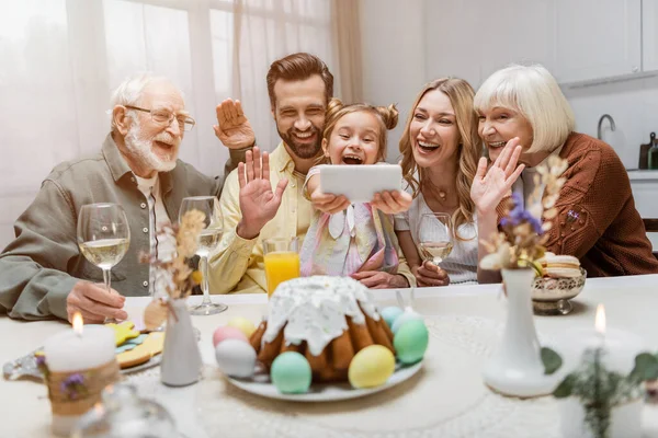 Cheerful family waving hands during video call on smartphone near table with easter dinner — Stock Photo