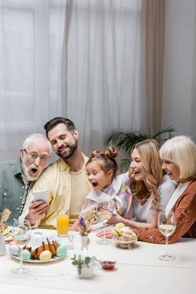 Uomo sorridente mostrando smartphone alla famiglia stupita durante la cena di Pasqua — Foto stock