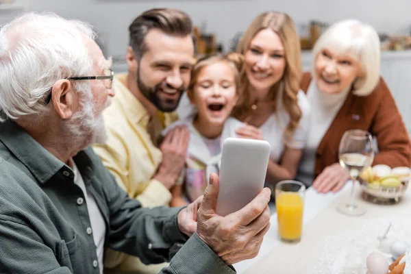 Aîné prenant selfie sur téléphone portable avec la famille floue pendant le dîner de Pâques — Photo de stock