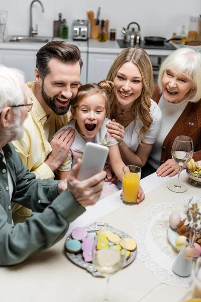 Senior man holding smartphone near excited girl with open mouth and happy family — Stock Photo