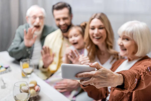 Senior woman taking selfie on smartphone with blurred family during easter celebration — Stock Photo