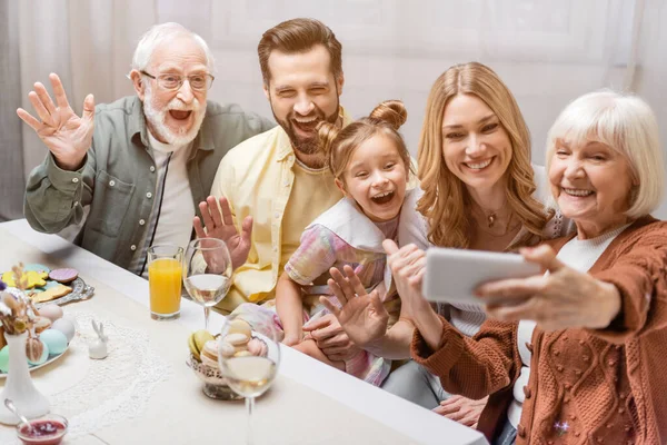 Mujer mayor sosteniendo teléfono inteligente cerca de la familia emocionada agitando las manos durante la cena de Pascua - foto de stock