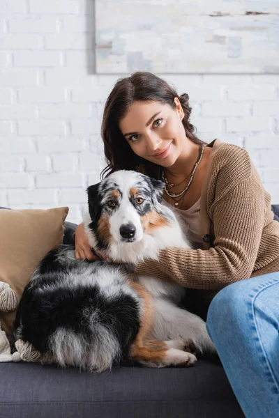 Pleased young woman cuddling australian shepherd dog while sitting on couch — Stock Photo