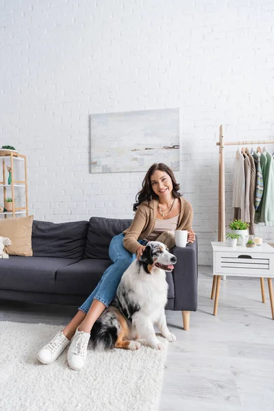 Happy young woman sitting on sofa and cuddling australian shepherd dog while holding cup of coffee — Stock Photo