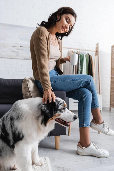 Smiling young woman cuddling australian shepherd dog and holding cup of coffee — Stock Photo