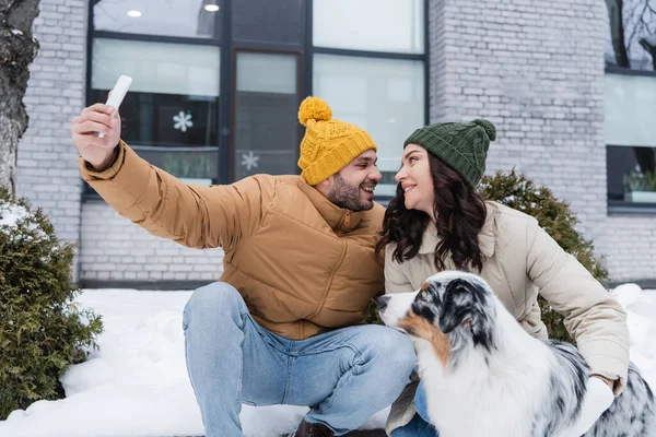 Happy man in knitted hat taking selfie with cheerful girlfriend and australian shepherd dog in winter — Stock Photo