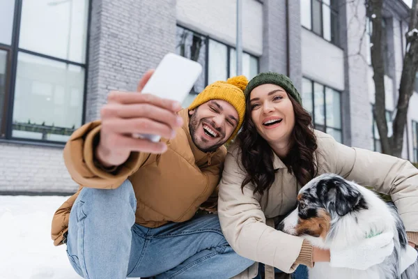 Smiling man in knitted hat taking selfie with girlfriend and australian shepherd dog in winter — Stock Photo