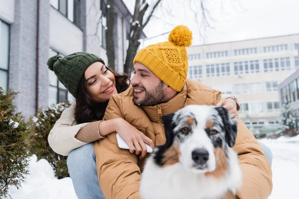 Smiling woman in knitted hat holding smartphone and hugging boyfriend near australian shepherd dog in winter — Stock Photo