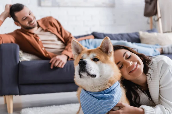 Smiling young woman hugging akita inu dog near blurred boyfriend on couch — Stock Photo