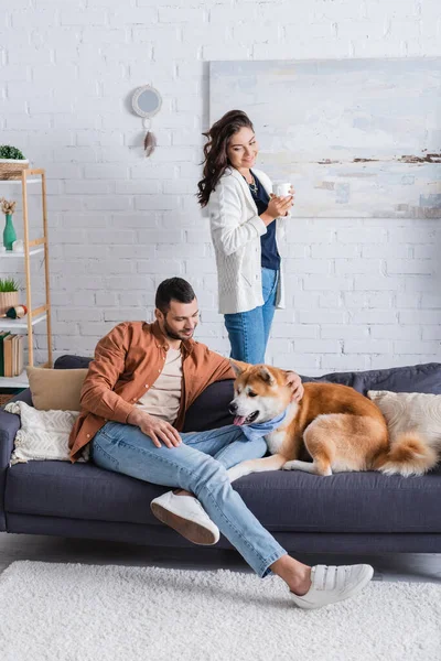 Bearded man sitting on couch and cuddling akita inu dog near girlfriend with cup — Stock Photo