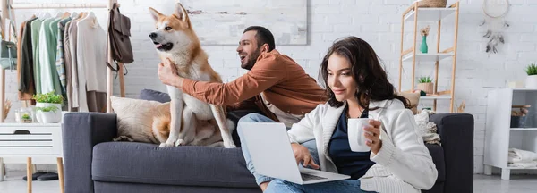 Young woman with cup using laptop near boyfriend cuddling akita inu dog on sofa, banner — Stock Photo
