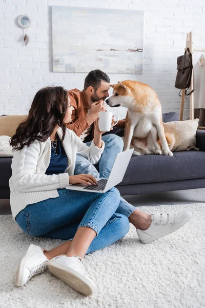 Smiling young woman with cup sitting on carpet and using laptop near boyfriend cuddling akita inu dog — Stock Photo