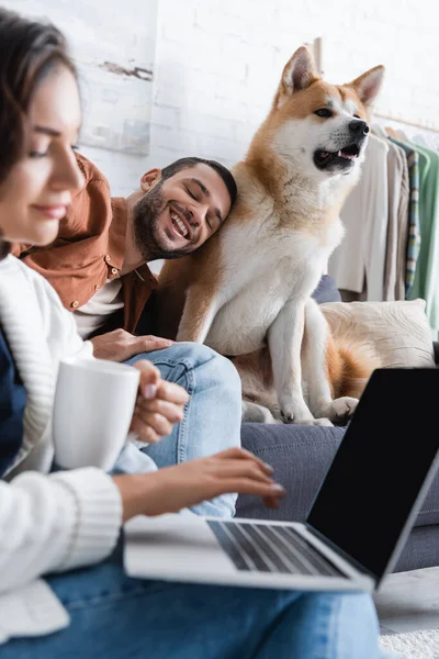Blurred young woman with cup using laptop near cheerful boyfriend cuddling akita inu dog — Stock Photo