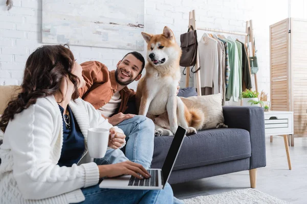 Young freelancer with cup using laptop near smiling boyfriend and akita inu dog on sofa — Stock Photo