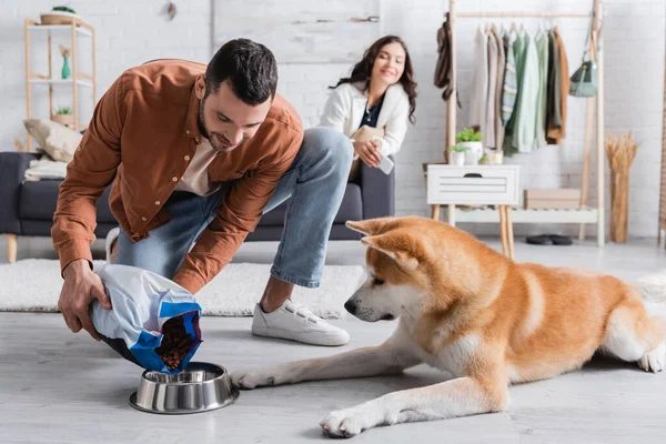Счастливый мужчина добавлял корм для домашних животных в миску рядом с собакой акита-ину и размытой подругой — стоковое фото
