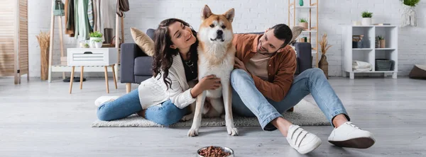 Felice giovane coppia che abbraccia akita inu cane vicino ciotola con cibo per animali domestici in soggiorno, banner — Foto stock