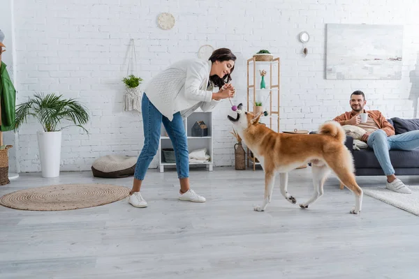 Happy young woman playing with akita inu dog near bearded boyfriend with cup of coffee sitting on couch — Stock Photo