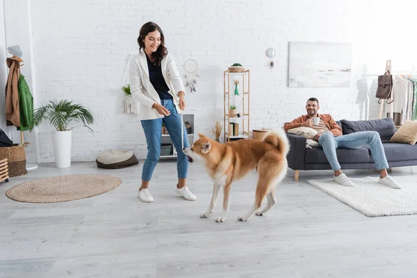 Smiling young woman playing with akita inu dog near happy boyfriend with cup of coffee sitting on couch — Stock Photo