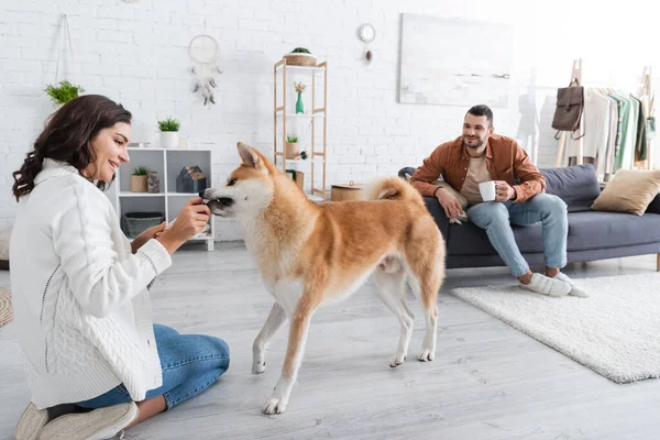 Happy young woman playing with akita inu dog near bearded boyfriend with cup of coffee — Stock Photo