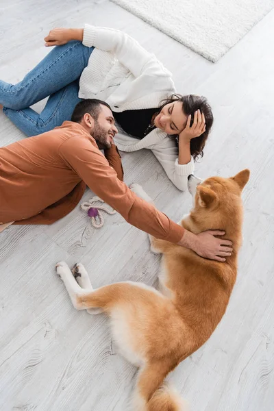 Top view of happy young couple looking at each other while lying of floor with akita inu dog — Stock Photo